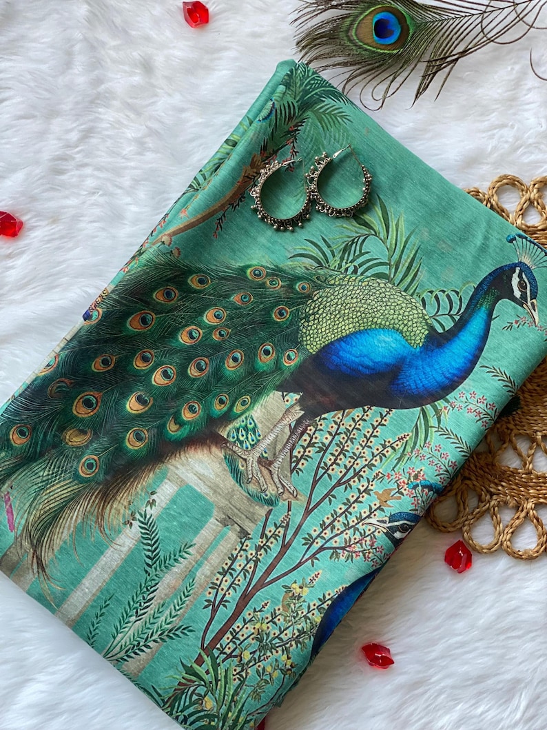 May include: A folded green fabric featuring a vibrant peacock print with blue and gold plumage, surrounded by botanical illustrations of leaves and branches. Two silver hoop earrings are placed on the fabric surface near the peacock.