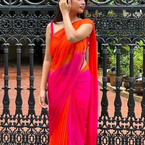 Może przedstawiać: Żywe sari ombre w odcieniach pomarańczu i różu. Materiał układa się elegancko, z kontrastującym różowym paskiem. Sari jest noszone w tradycyjnym stylu, tworząc płynną sylwetkę. Tło obejmuje czarną kutą bramę i zieleń.