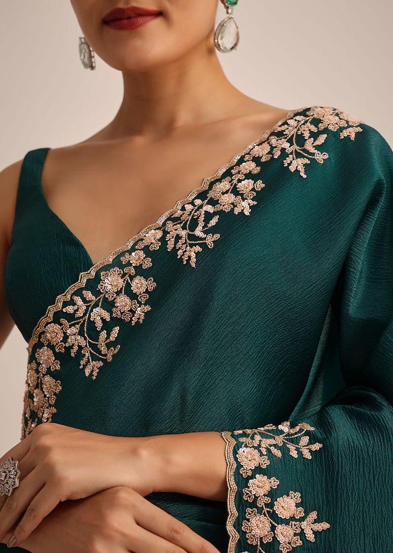 May include: A deep emerald green sleeveless blouse featuring a V-neckline and a textured fabric. The garment is adorned with intricate light gold floral embroidery and sequin detailing along the neckline and the sleeve edge.