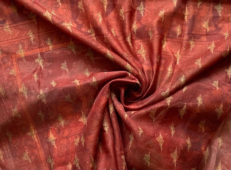 May include: A deep red fabric featuring a repeating gold-colored pattern of small, stylized floral or leaf-like motifs. The material has a translucent, lightweight quality with visible folds and gathers that create a swirling, textured appearance.