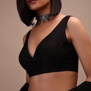 Puede incluir: Una blusa negra sin mangas con un escote en V profundo y costuras estructuradas a lo largo del corpiño. La prenda se combina con un collar tipo gargantilla de metal plateado con intrincados detalles grabados y pequeños elementos colgantes.