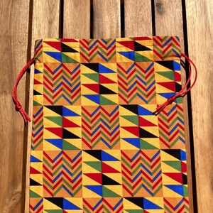 Pode incluir: Uma bolsa retangular com cordão com um padrão geométrico vibrante em amarelo, vermelho, azul, verde e preto. A bolsa tem um fecho de cordão vermelho. O padrão apresenta ziguezagues e triângulos.