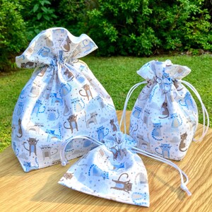Könnte beinhalten: Drei weiße Kordelzugbeutel mit einem blauen und braunen Katzendruck. Die Beutel sind in verschiedenen Größen und liegen auf einer Holzoberfläche.