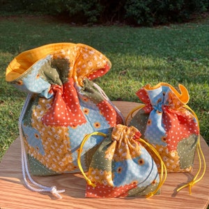 Peut inclure: Trois sacs en tissu &agrave; cordon de diff&eacute;rentes tailles avec un motif patchwork compos&eacute; de motifs floraux orange, jaune, bleu et vert.