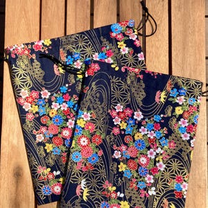 Pode incluir: Duas sacolas azul marinho com cordão com um vibrante padrão floral em vermelho, rosa, amarelo e azul. O design inclui detalhes dourados e um fecho de cordão preto. As sacolas são feitas de tecido e são exibidas em uma superfície de madeira.