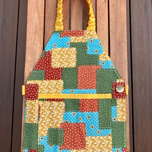 Puede incluir: Un delantal de patchwork con una correa para el cuello y un bolsillo amarillos. El delantal presenta una variedad de cuadrados de tela coloridos en azul, verde, rojo y amarillo, con estampados florales y de lunares. Se ve un cierre de botón en el lateral.