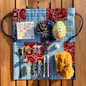 Op de afbeelding: Een patchwork stoffen tas met een blauw trekkoord, met een mix van rode paisley, blauwe gingham en patronen. De tas bevat een verscheidenheid aan kleine items, waaronder een dennenappel, een beschilderd ei, een pop, een lepel en een spons.