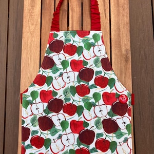 May include: A white apron with a red apple pattern and green leaves. The apron has a red neck strap and a red button on the side. The apron is made of fabric and is designed for kitchen use.