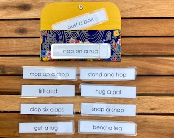 May include: A set of ten clear plastic cards with black text on a wooden surface. The cards have phrases like "dust a box", "nap on a rug", "mop up a drop", "stand and hop", "lift a lid", "hug a pal", "clap six claps", "snap a snap", "get a rug", and "bend a leg".