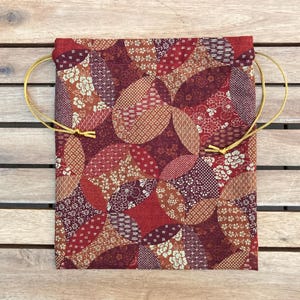 May include: A rectangular fabric drawstring pouch featuring a repeating geometric pattern of overlapping circles in shades of deep red, burgundy, and burnt orange, accented with small floral and textured motifs and thin gold-colored cord ties.