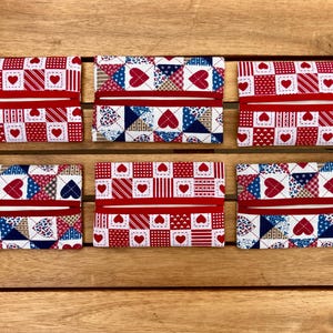 May include: Six fabric tissue holders with red, white, and blue patterns. The designs include hearts, stripes, and patchwork squares. Each holder has a red band across the middle. The holders are arranged on a wooden surface.