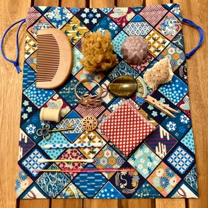 Pode incluir: Uma bolsa de cordão azul e dourada cheia de vários itens, incluindo um pente de madeira, uma esponja natural, um pequeno sabonete roxo, uma concha, uma pedra olho de tigre, um conjunto de anéis de ouro, uma bobina de fio, um pequeno botão de madeira, um pano estampado vermelho e branco, um par de prendedores de roupa de madeira e dois cotonetes.