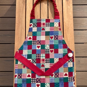 Puede incluir: Un delantal de patchwork con un diseño de corazón rojo. El delantal presenta una variedad de cuadrados coloridos en tonos de azul, verde, rojo y blanco. Tiene una correa roja y una correa para el cuello roja. Un botón blanco está en el lateral.