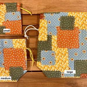 Peut inclure: Trois sacs &agrave; cordon en tissu patchwork de diff&eacute;rentes tailles&nbsp;: petit, moyen et grand. Les sacs sont faits de diff&eacute;rents tissus dans des tons de bleu, vert, jaune et orange. Les sacs sont tous attach&eacute;s avec des cordons de serrage blancs.