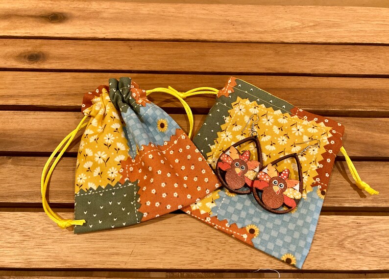 Peut inclure: Une paire de boucles d'oreilles en bois en forme de dinde avec un motif peint brun, orange et jaune. Les boucles d'oreilles sont dans un petit sac en tissu patchwork avec des cordons jaunes.