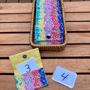 May include: A collection of small fabric pouches featuring a vibrant floral pattern in yellow, pink, red, and blue. One open pouch displays a white card with the number 3, while a separate clear plastic case holds a card with the number 4.