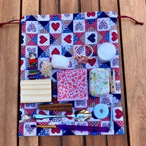 Pode incluir: Uma bolsa de tecido patchwork vermelho, branco e azul cheia de vários itens pequenos, incluindo um bloco de madeira, uma boneca pequena, uma vela, um anel de metal, uma bola de algodão, uma lata pequena, um pau de madeira, uma colher de metal, um disco de metal e uma lixa de unha roxa.
