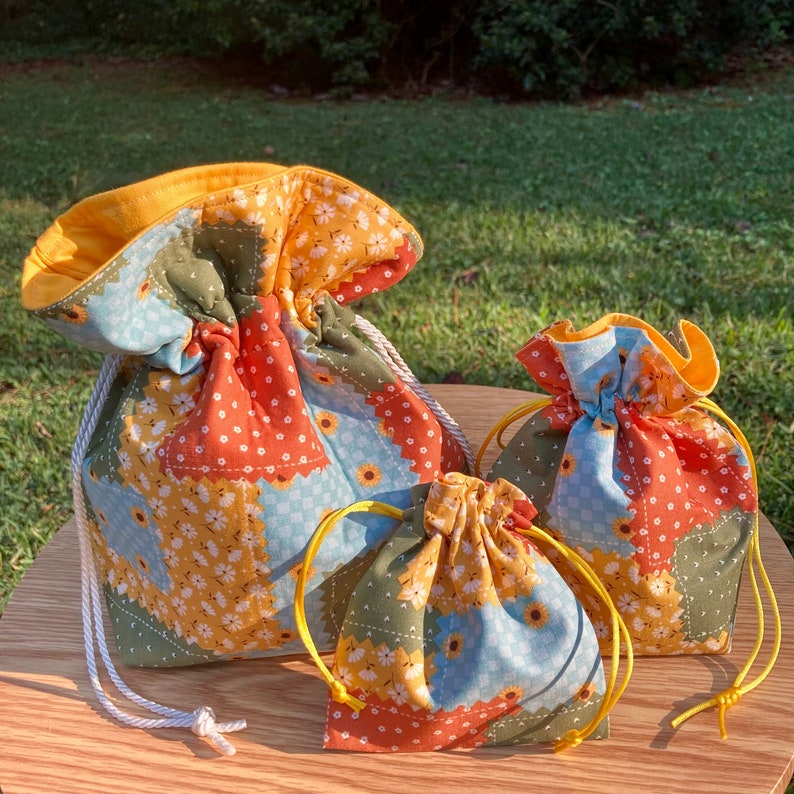 Peut inclure: Trois sacs en tissu &agrave; cordon de diff&eacute;rentes tailles avec un motif patchwork compos&eacute; de motifs floraux orange, jaune, bleu et vert.