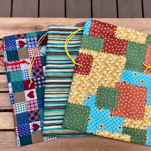 May include: Three rectangular fabric drawstring bags. The left bag features a blue, red, and green patchwork pattern with heart motifs. The center bag has horizontal brown, green, and blue stripes. The right bag displays a floral patchwork design.