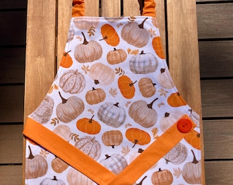 May include: A white apron featuring a repeating pattern of pumpkins in shades of orange, beige, and gray. The apron has an orange elastic neck strap and a V-shaped orange fabric trim across the front, accented with a single orange button on the right.