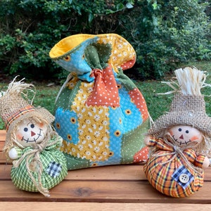 Peut inclure: Deux petits &eacute;pouvantails faits &agrave; la main avec des chapeaux en toile de jute et des corps en tissu patchwork. Les &eacute;pouvantails sont assis sur une surface en bois devant un sac &agrave; cordon en tissu patchwork.