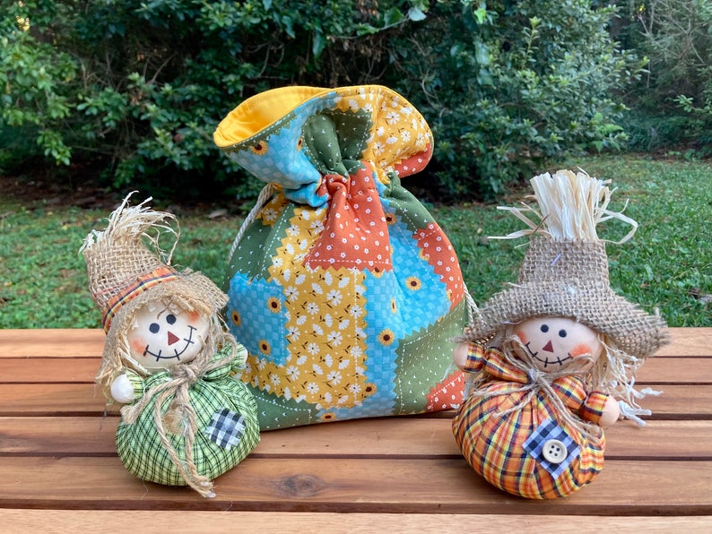 Peut inclure: Deux petits &eacute;pouvantails faits &agrave; la main avec des chapeaux en toile de jute et des corps en tissu patchwork. Les &eacute;pouvantails sont assis sur une surface en bois devant un sac &agrave; cordon en tissu patchwork.