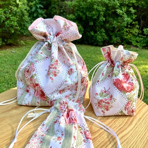 Pode incluir: Três sacos de cordão feitos de tecido branco com um design de patchwork com padrões florais rosa e verdes. Os sacos são de tamanhos diferentes e estão dispostos sobre uma superfície de madeira.