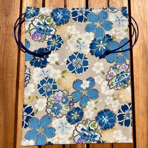 Pode incluir: Uma bolsa com cordão de tecido com um padrão floral. A bolsa apresenta um fundo cinza claro com um padrão repetido de flores azuis, brancas e roxas. Possui um cordão azul escuro. A bolsa é feita de tecido.