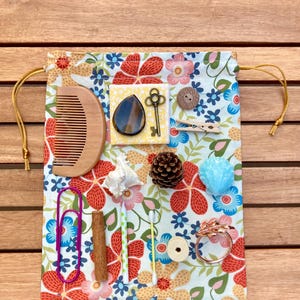 May include: A drawstring bag with a floral pattern, featuring a collection of small objects. These include a wooden comb, a key, a button, a paperclip, a pinecone, and a ring. The bag's design incorporates red and blue floral elements.