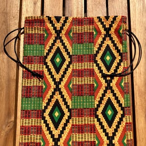 Pode incluir: Uma bolsa quadrada com cordão com um padrão vibrante de inspiração africana. O design apresenta formas geométricas repetidas em vermelho, amarelo, verde e preto. A bolsa tem cordões pretos para fechar e é feita de tecido.