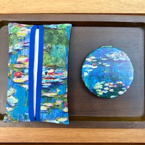 May include: A tissue box cover and a compact mirror featuring a water lilies painting in shades of blue, green, and red. The tissue box cover is rectangular with a blue band, and the mirror is round. Both items are on a dark brown tray.