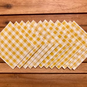 May include: A set of ten square cloth napkins arranged on a wooden surface. Each napkin features a yellow and white checkered pattern. The napkins are neatly stacked, showcasing their design and texture.