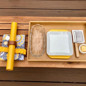 May include: A wooden tray with a yellow and gray patterned fabric, a yellow candle, a glass dish, a white plate with a yellow rim, a small metal grater, a wooden handle, and a small container with a yellow lid.