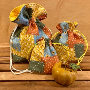 Peut inclure: Trois sacs &agrave; cordon en tissu patchwork dans des tons de bleu, vert, jaune et orange. Les sacs sont pos&eacute;s sur une surface en bois avec une petite citrouille en c&eacute;ramique jaune devant eux.