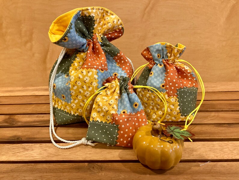 Peut inclure: Trois sacs &agrave; cordon en tissu patchwork dans des tons de bleu, vert, jaune et orange. Les sacs sont pos&eacute;s sur une surface en bois avec une petite citrouille en c&eacute;ramique jaune devant eux.