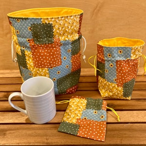 Peut inclure: Trois sacs en tissu avec un motif patchwork dans des tons de bleu, vert, orange et jaune. Les sacs ont des cordons de serrage et sont faits d'un tissu matelass&eacute; doux. Une tasse en c&eacute;ramique blanche est &eacute;galement incluse dans l'image.