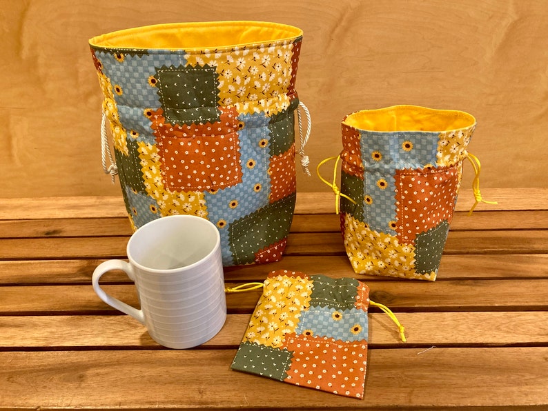 Peut inclure: Trois sacs en tissu avec un motif patchwork dans des tons de bleu, vert, orange et jaune. Les sacs ont des cordons de serrage et sont faits d'un tissu matelass&eacute; doux. Une tasse en c&eacute;ramique blanche est &eacute;galement incluse dans l'image.