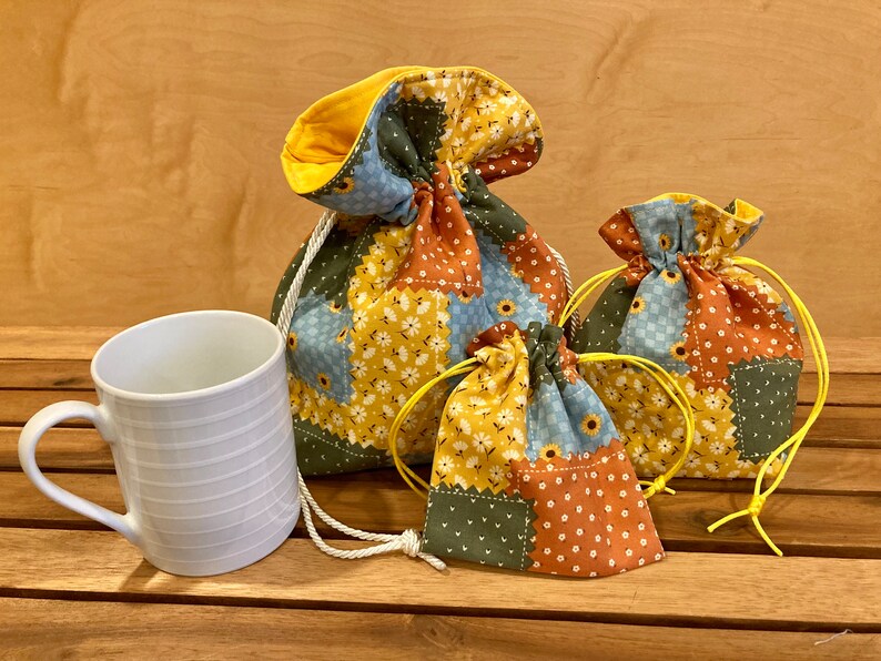 Peut inclure: Trois sacs en tissu avec cordon de serrage de diff&eacute;rentes tailles, avec un motif patchwork de carr&eacute;s jaunes, bleus, verts et oranges. Les sacs sont en tissu de coton et ont un cordon de serrage jaune.