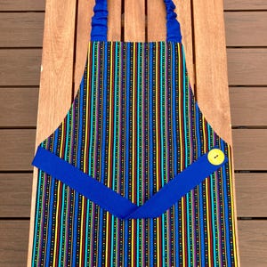 May include: A kitchen apron featuring a vertical striped pattern in black, blue, yellow, red, and turquoise. The apron includes a solid blue elasticized neck strap and a matching blue waist tie secured with a single bright yellow button.