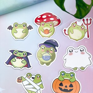 May include: A collection of Halloween-themed stickers featuring cartoon frogs. Each sticker depicts a frog in a costume, including a bat, devil, vampire, witch, mummy, and ghost. The stickers are colorful with a white border.