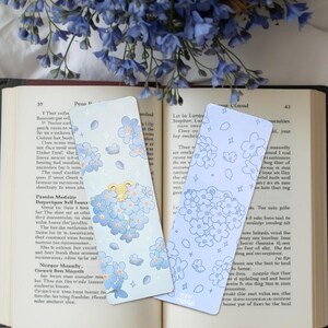 Peut inclure: Deux marque-pages floraux avec des motifs de fleurs bleues sont présentés sur un livre ouvert. L'un des marque-pages présente un personnage de grenouille entouré de fleurs, tandis que l'autre a un motif floral bleu clair. Les marque-pages sont rectangulaires.