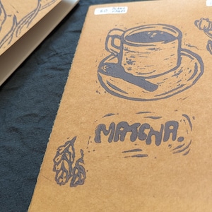 May include: A brown notebook featuring a coffee cup and saucer illustration, with the word "MASCHA" printed beneath. Another notebook with a bird illustration is partly visible. The illustrations are in dark blue ink.