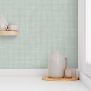 K&ouml;nnte beinhalten: Ein hellgr&uuml;ner, strukturierter Tapetenhintergrund. Ein wei&szlig;er Keramikkrug und eine kleine Tasse stehen auf einem Holztablett. Ein kleines Holzregal h&auml;lt zwei kleine Tassen und eine wei&szlig;e Keramikvase.