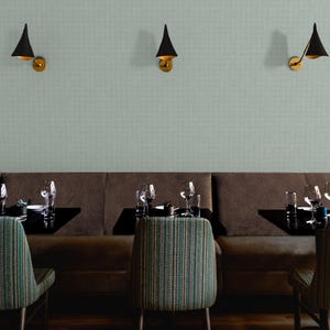 K&ouml;nnte beinhalten: Ein Restaurantinterieur mit einer braunen Sitzbank, Tischen und St&uuml;hlen mit gestreifter Polsterung. Schwarze, kegelf&ouml;rmige Wandleuchten mit goldenen Akzenten sind an einer hellgr&uuml;nen, strukturierten Wand angebracht. Weingl&auml;ser und Gedecke auf den Tischen.