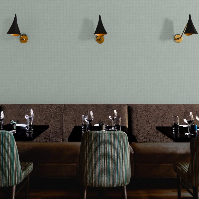 K&ouml;nnte beinhalten: Ein Restaurantinterieur mit einer braunen Sitzbank, Tischen und St&uuml;hlen mit gestreifter Polsterung. Schwarze, kegelf&ouml;rmige Wandleuchten mit goldenen Akzenten sind an einer hellgr&uuml;nen, strukturierten Wand angebracht. Weingl&auml;ser und Gedecke auf den Tischen.