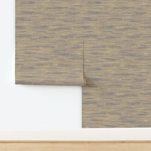 May include: Neutral-toned wallpaper with a woven texture, featuring horizontal streaks of blue and tan. The wallpaper is partially unrolled, revealing the backing. The bottom of the image shows a white baseboard and a wooden floor.