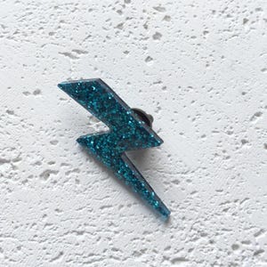 Pode incluir: Um broche de raio azul turquesa com glitter. O broche é feito de plástico e tem um suporte de metal.