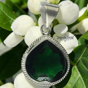 Puede incluir: Un colgante en forma de corazón con una piedra preciosa facetada de color verde intenso engastada en un marco de plata con un borde de cuerda trenzada. El colgante incluye una anilla de plata en la parte superior. El texto DsgnsbyDavid.com es visible.