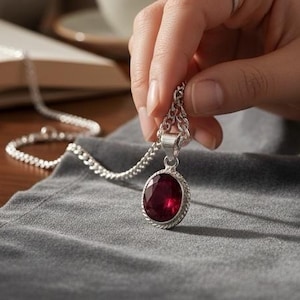 Op de afbeelding: Een zilveren hangerketting met een grote, ovale, rode edelsteen. De ketting wordt door een hand vastgehouden, met een boek, koffiekopje en kleine plant op de achtergrond. De tekst "NATURAL BERYL BIXBITE RED EMERALD" staat bovenaan.