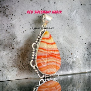 May include: A silver pendant with a red and white striped agate stone. The stone is shaped like a teardrop and has a silver bail. The pendant is on a gray background. The text "RED SULEMANI HAKIK" is above the pendant. The text "DsgnByDavid.com" is below the pendant.